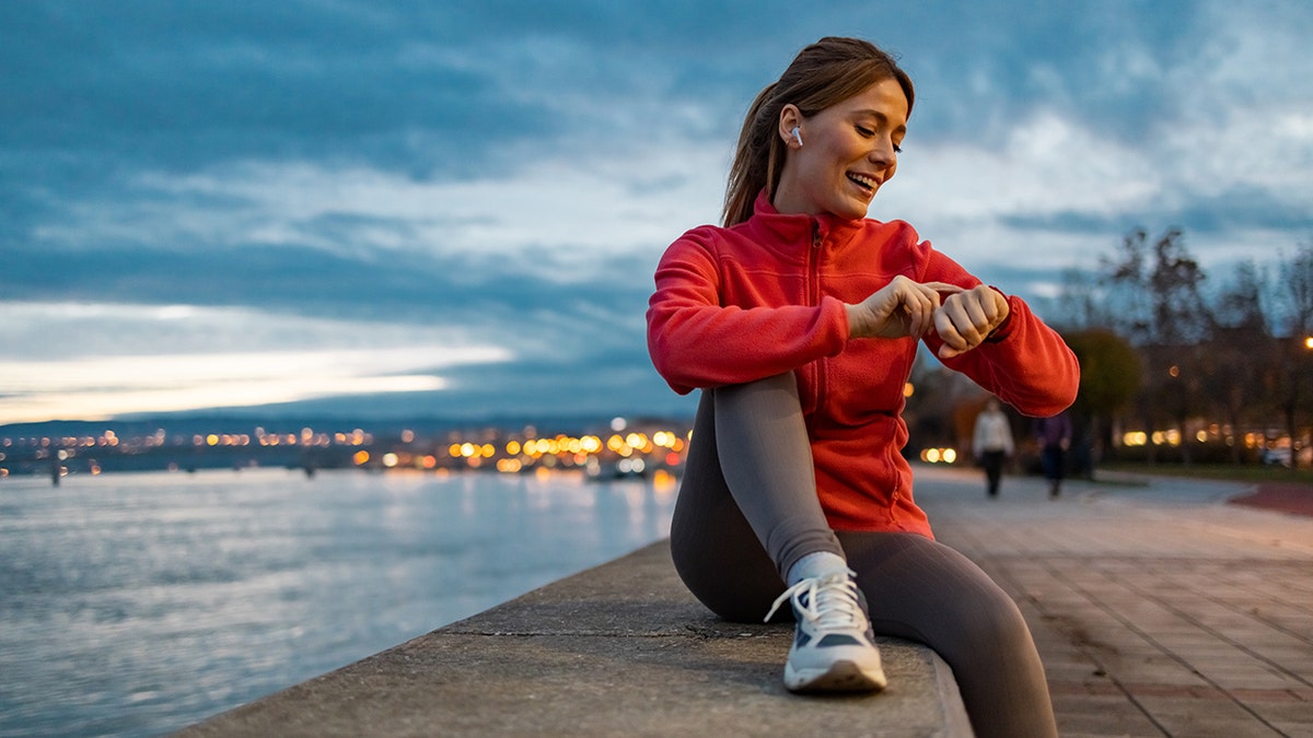 Woman in running gear checks her heart rate on a smart watch. She is wearing a red long sleeve quarter zip and sits along the edge of the water.