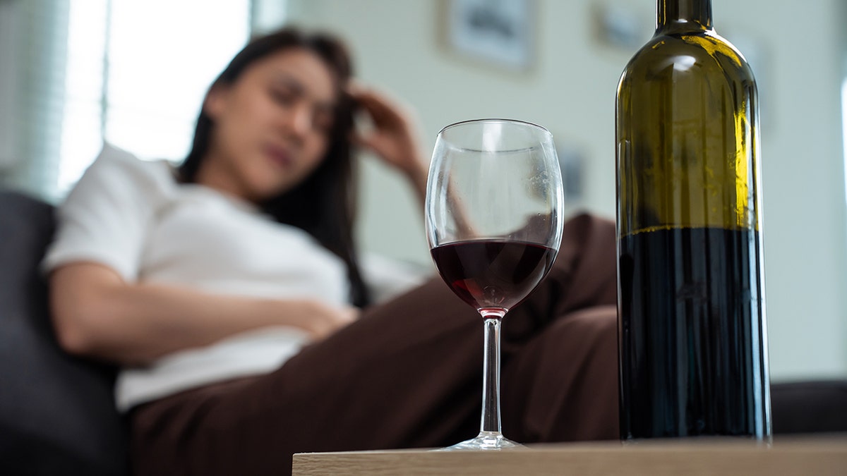 Woman is blurred in background and holding her head while sitting on a couch and ooking towards the wine bottle and glass of red wine in the foreground.
