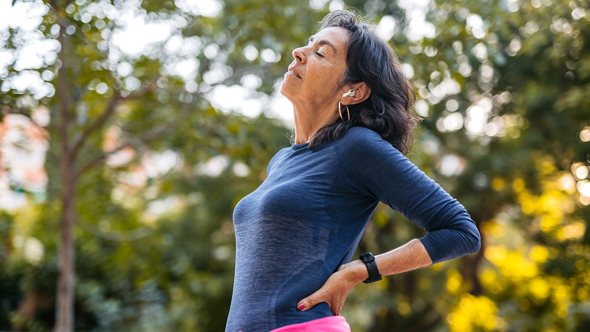 Older woman outside, stretching her back and closing her eyes with her face angled up towards the sun. She has long greying hair and is wearing a watch and exercise clothes in a dark blue material.