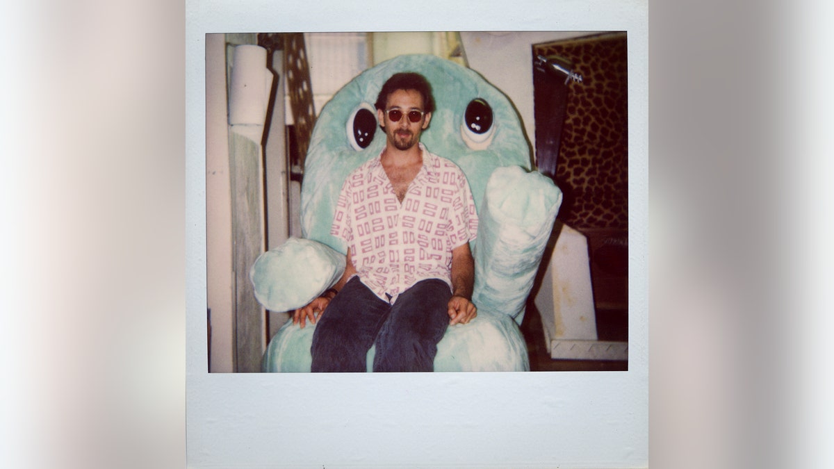 Paul Reubens wearing a pink plaid shirt sitting on a blue plush bear.