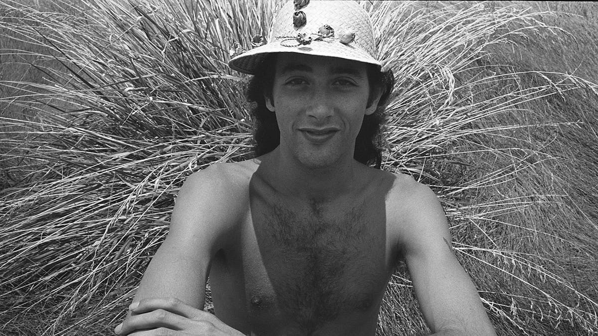 A young Paul Reubens wearing a hat and sitting in front of grass.