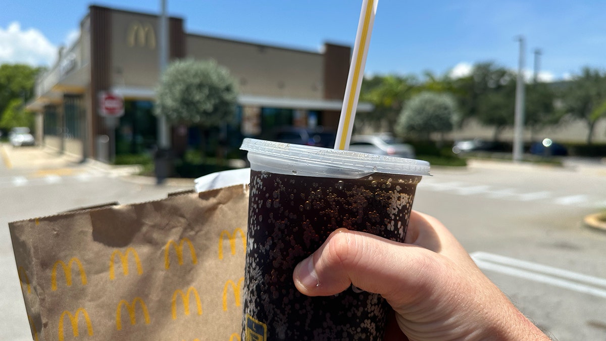 A man's hand holds a McDonald's bag and a large soda with a plastic straw outside of the fast-food restaurant.