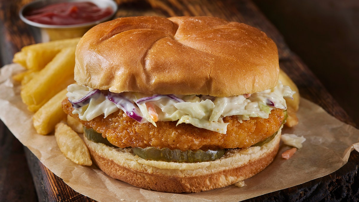 A hot chicken sandwich on a seedless bun with pickles, cole slaw and a side of fries is shown.