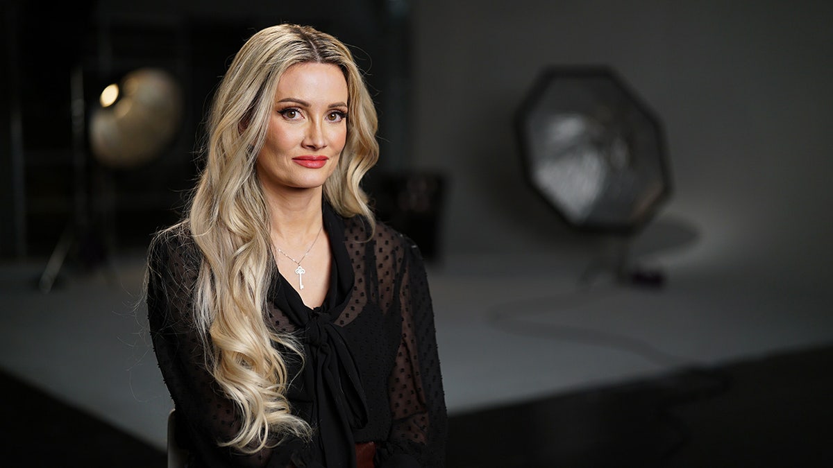 Holly Madison smirking in a black sheer top inside a studio.