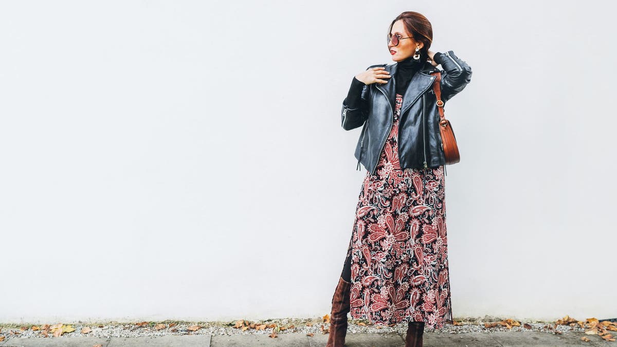 A female in Boho's clothes smiled similar to a long colored dress with a black leather motorcycle jacket with a brown leather flap that pretends against the background of the white wall.
