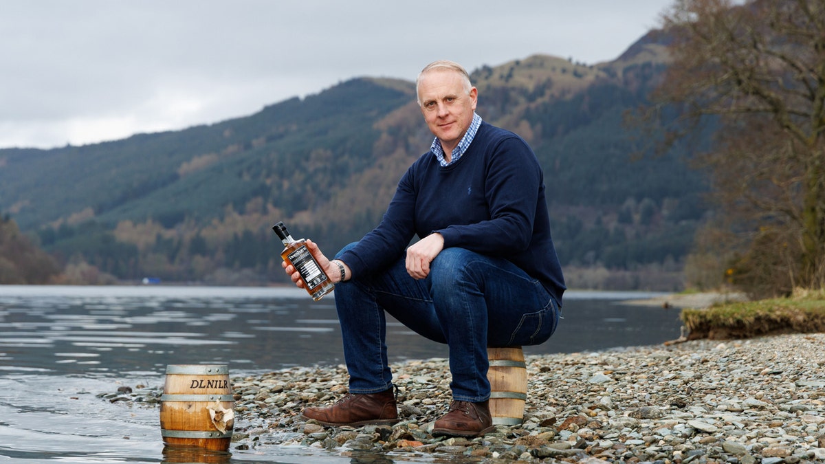 Domhayn founder James Patterson poses with a spirit bottle that was submerged in Loch Ness. He sits on one of the casks that was submerged while holding a bottle with Loch Ness as his backdrop.