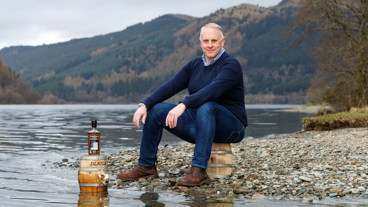 Domhayn founder James Patterson poses with a spirit bottle that was submerged in Loch Ness. He sits on one of the casks that was submerged while standing with Loch Ness as his backdrop.