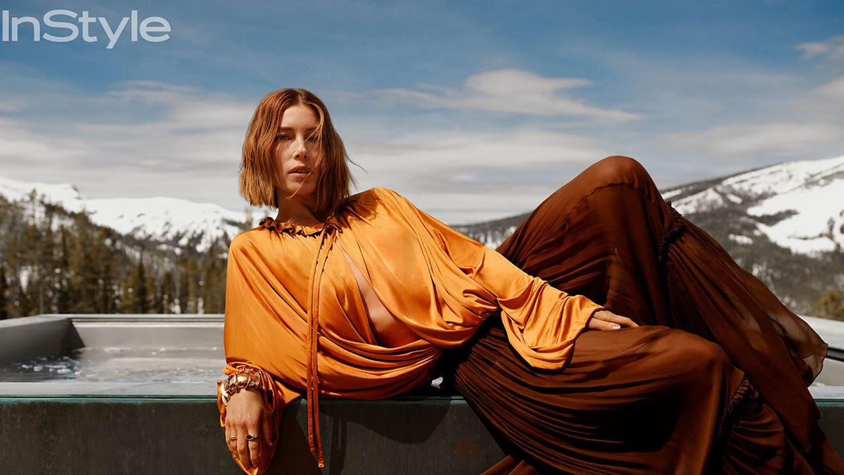 jessica biel lounging in a orange top and maroon skirt in front of snow-covered mountains