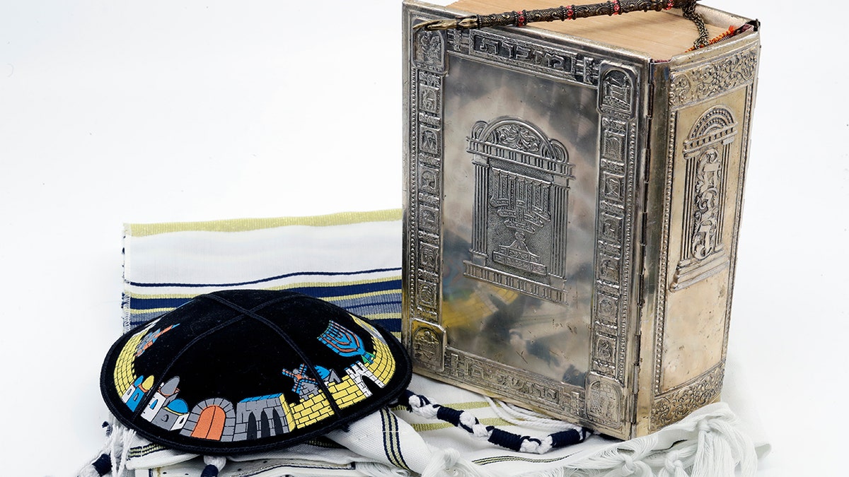 silver hardcover Jewish Torah on top of a folded tallit beside a black decorated yarmukle