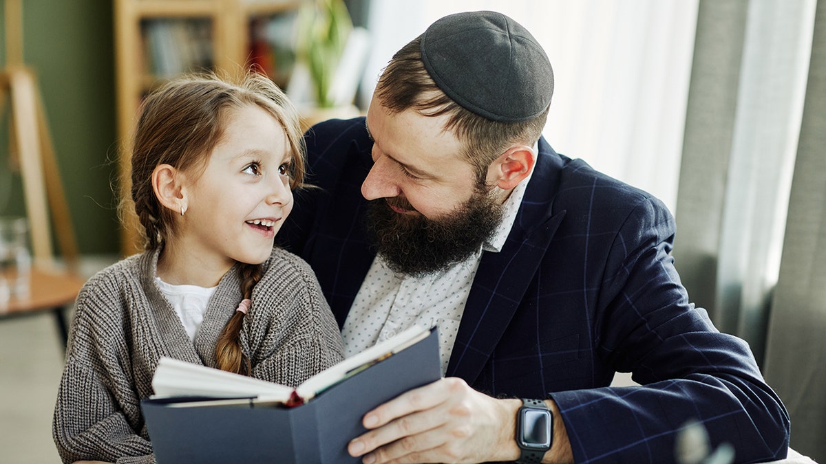 Portrait of smiling Jewish father reading book with daughter at home. The father wears a yarmukle as he looks at his daughter sat on his lap as she gazes at him with a smile and bright eyes