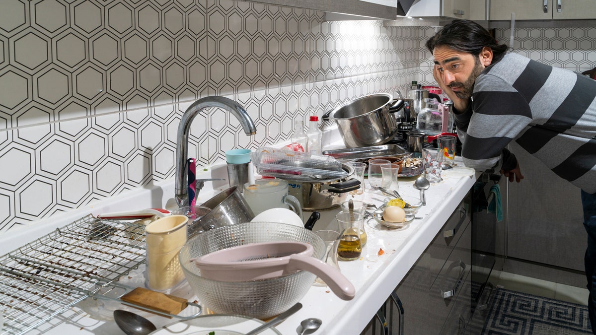 A bearded man stares at dirty utensils, pots and dishes in a kitchen.