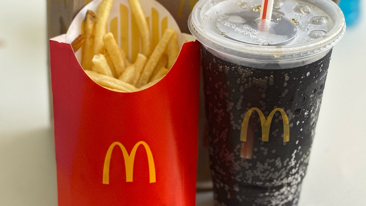 An order of large fries and a large soda from McDonald's are propped in front of a McDonald's bag on a table.