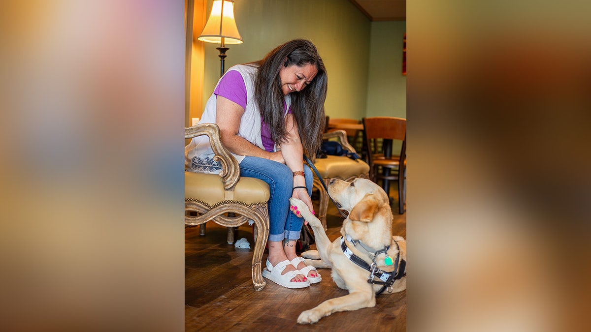 A woman smiles as she receives a paw shake from her guide dog in a warmly lit room
