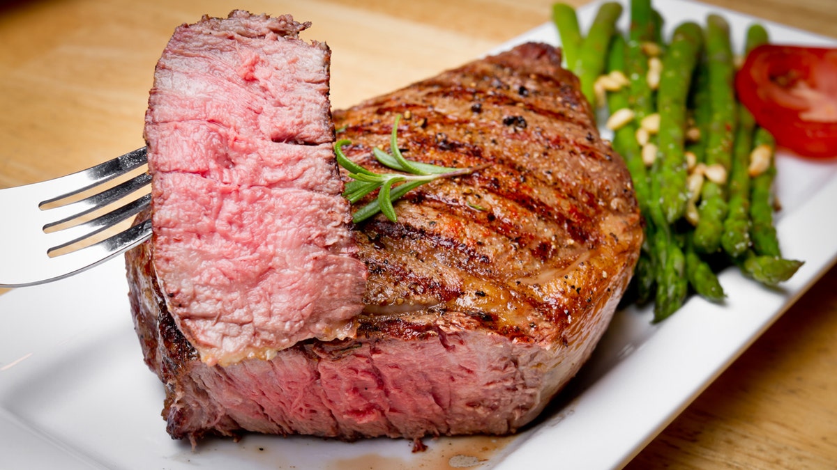 A big ribeye steak cooked medium rare with a bite cut out and speared on a fork with asparagus and tomato on the side. The meal is presented on a square white plate on a light wooden table. The steak also has a sprig of rosemary and perfect grill marks.