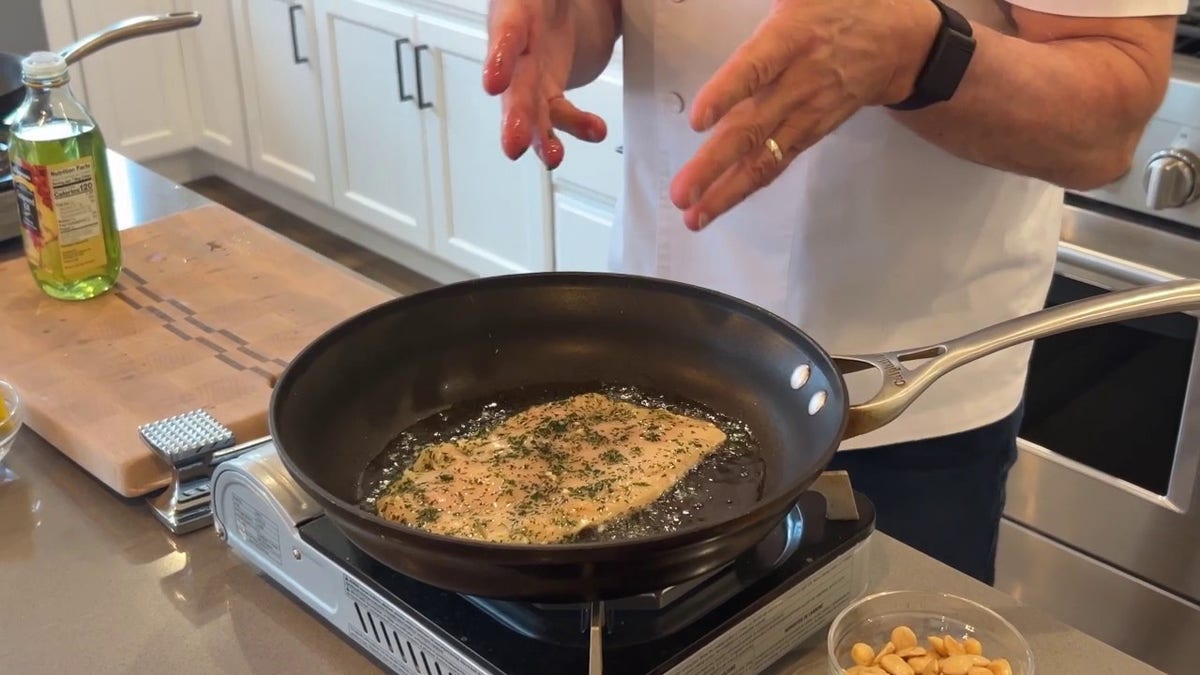 Robert Irvine points to chicken paillard cooking in a pan with grapeseed oil.