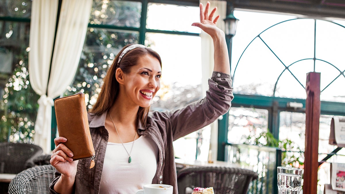 A smiling woman seated at a table at a restaurant raises her hand that she's ready to pay her bill.