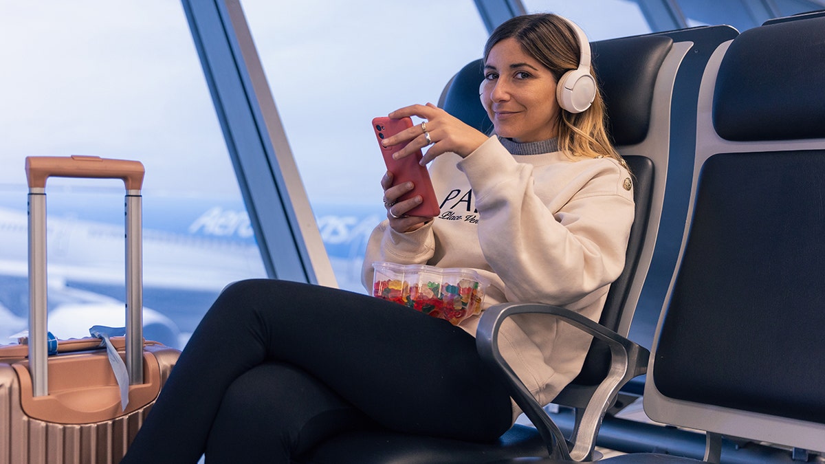 A woman wearing headphones smiles while holding her phone with gummy bears in her lap as she sits at an airport next to her luggage.