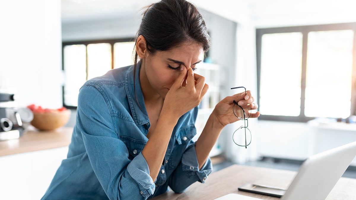 Woman with denim shirt pinching her nose in stress while holding vessels in front of a laptop at home.