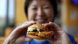An Asian woman, whose face is out of focus, holds a chicken sandwich.