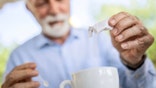 Older man pouring sugar into a cup of coffee, only the sugar and cup are in focus.