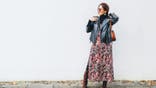 Smiling female dressed boho fashion style colorful long dress with black leather biker jacket with brown leather flap bag posing on the white wall background.