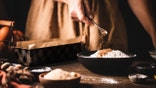 A woman preparing banana bread adds cinnamon in a wheat flour bowl over a kitchen counter.