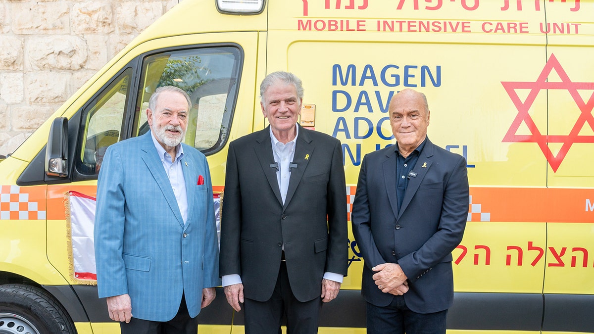 Franklin Graham, Mike Huckabee, and Eli Bin stand in front of a yellow Magen David Adom armored ambulance in Jerusalem