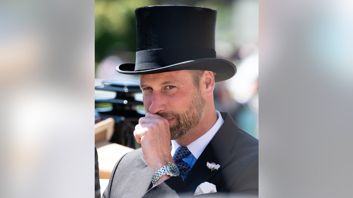 Prince William with his fist to his face as he smirks with a dark suit and a black top hat.