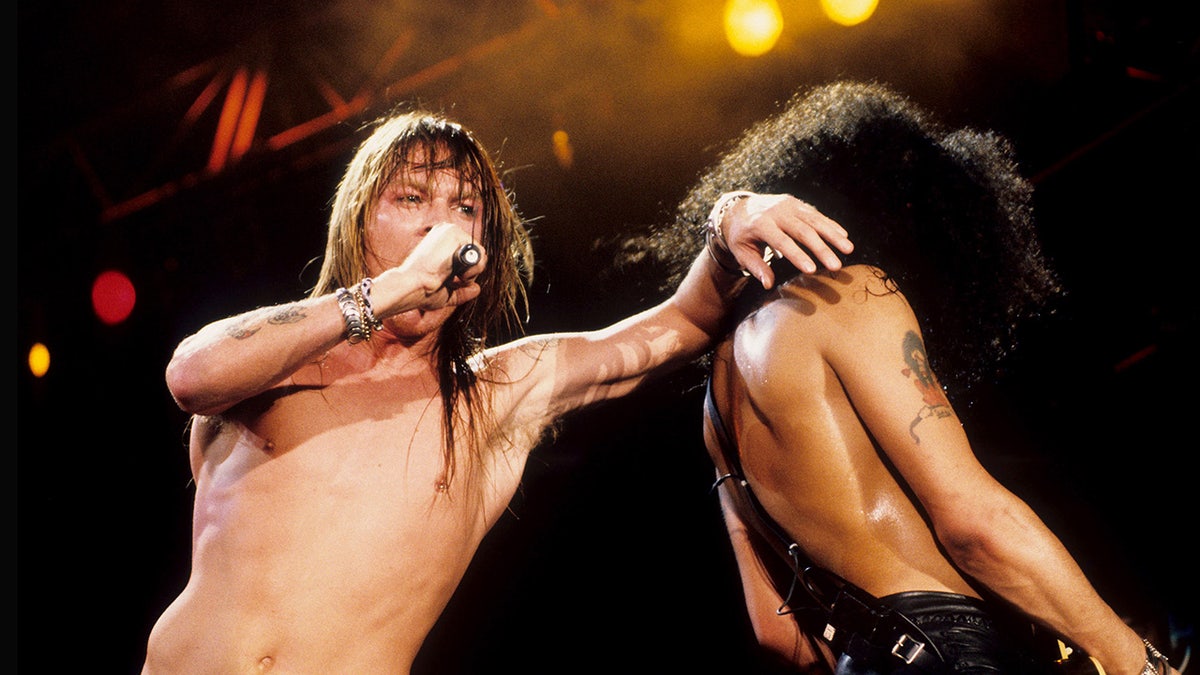 Axl Rose leaning connected Slash while singing connected stage.