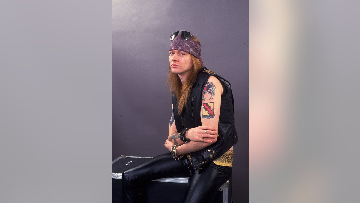 Axl Rose wearing leather pants and a achromatic vest pinch a purple bandana showing disconnected his tattoos.