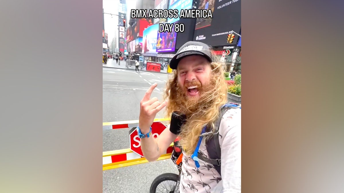 US comedian Sebastian 'Seabass' Fowler taking a selfie in Times Square with a big smile on his face with a caption acorss the top of the photo that reads "BMX across america day 80"