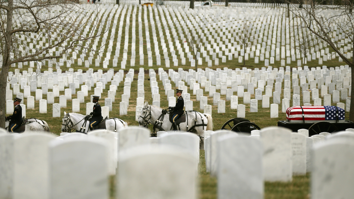A military funeral procession at Arlington National Cemetery features a flag-draped casket carried on a horse-drawn caisson, with uniformed service members on horseback riding past rows of white marble headstones.