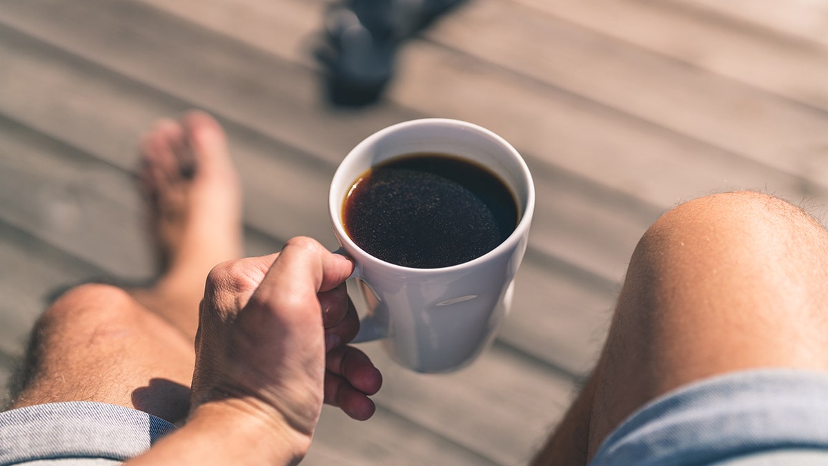 A man is barefoot and holding a white mug filled with coffee.