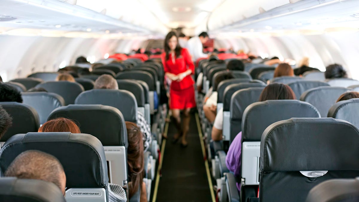 flight attendant walks down the aisle of a plane, as passengers are seated facing forward