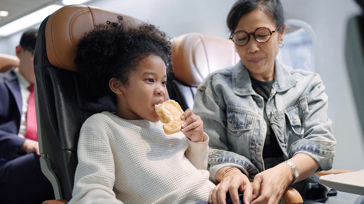Mother and preteen daughter sitting in passenger airplane enjoy eating donut. Mother helping to adjust and tight seatbelt on an airplane for safe flight and enjoying travelling