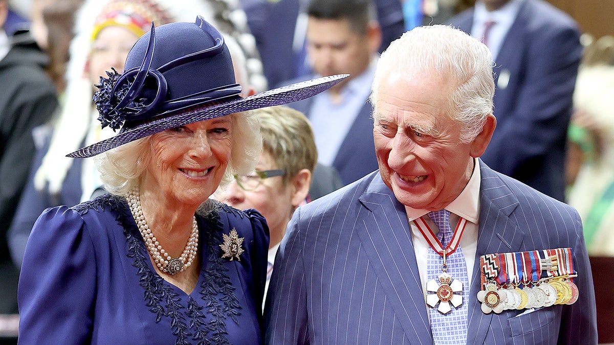 King Charles in a blue suit with medals smiling at Queen Camilla in a blue dress and matching hat.