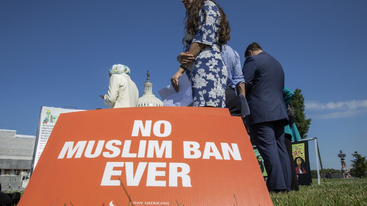Activists gather on a lawn near the U.S. Capitol, with an orange sign in the foreground reading