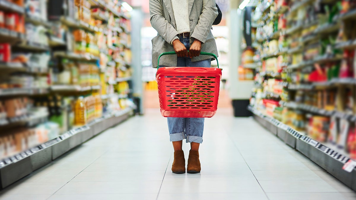 A customer's legs and their grocery basket are seen standing in the middle of a grocery store aisle.