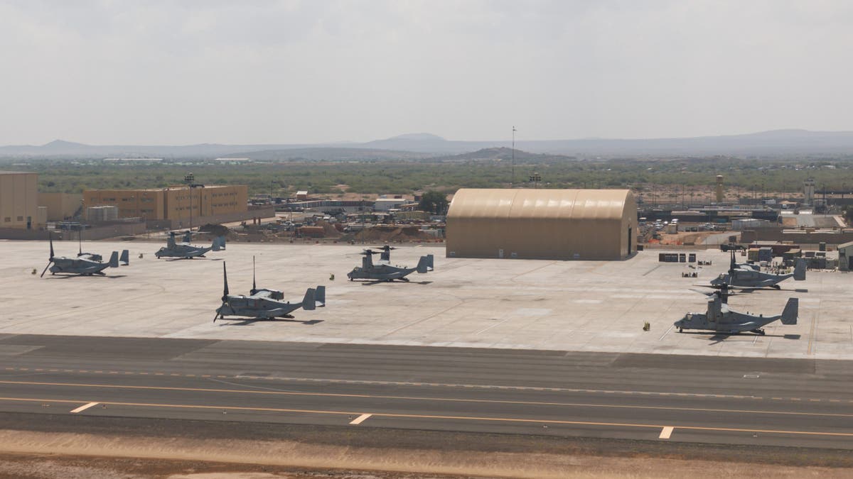 U.S. military aircraft, including MV-22 Ospreys, are parked on the tarmac at a forward operating base in Djibouti, with aircraft hangars and support structures visible under hazy skies near the Horn of Africa.