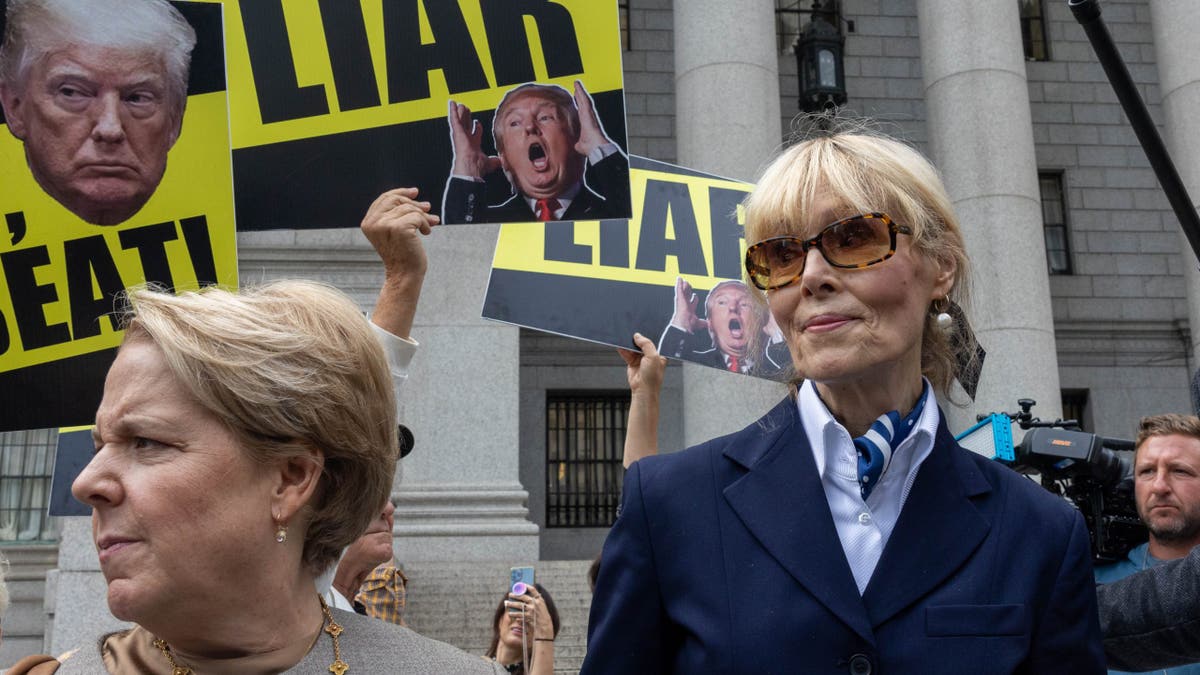 E. Jean Carroll exits a New York courthouse alongside her attorney, with protesters in the background holding signs that read