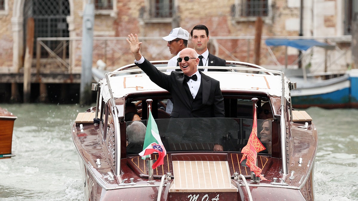 Jeff Bezos waving on a water taxi in a black tuxedo