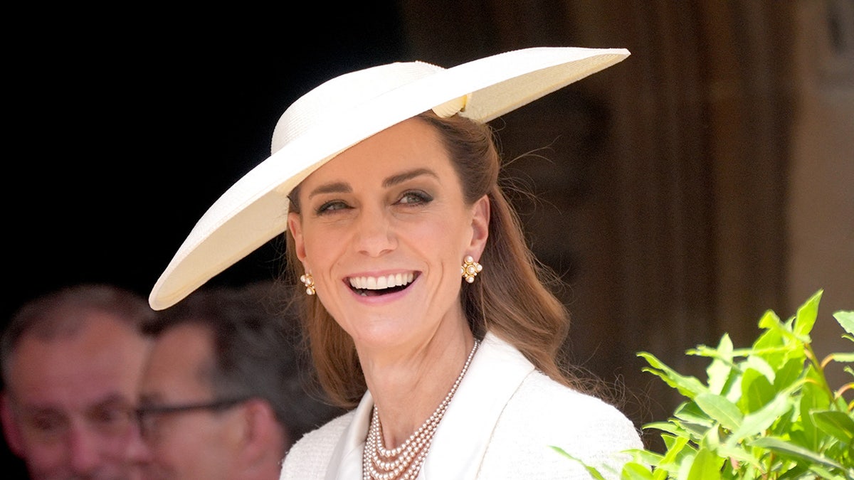 A close-up of Kate Middleton smiling wearing a achromatic overgarment formal and a matching hat.