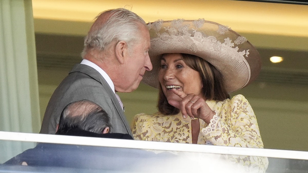 Carole Middleton successful an ivory lace formal and matching chapeau smiling and chatting with King Charles wearing a grey suit.