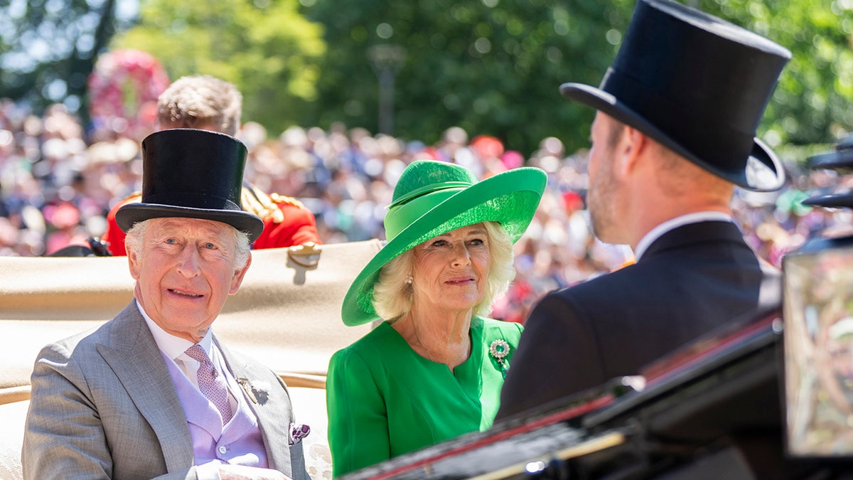 King Charles and Queen Camilla wearing ceremonial suits and festive hats sitting crossed from Prince William connected a royal carriage.