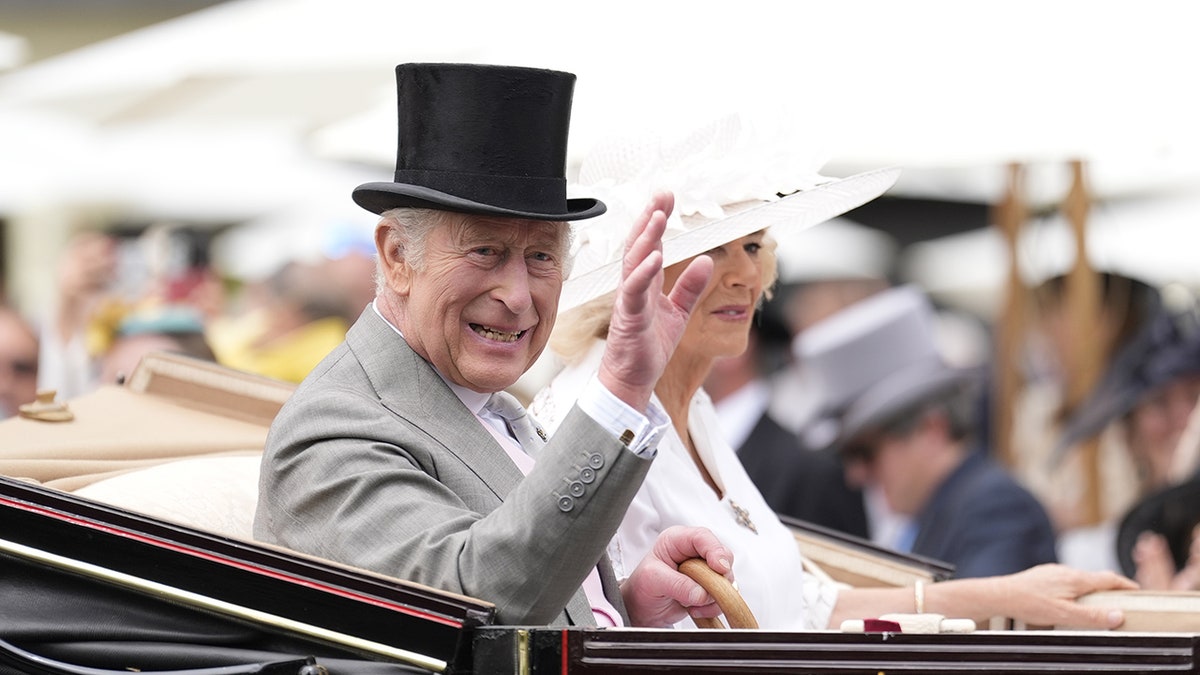 King Charles in a grey suit and black top hat waving in a royal carriage next to Camilla wearing all white with a matching hat.