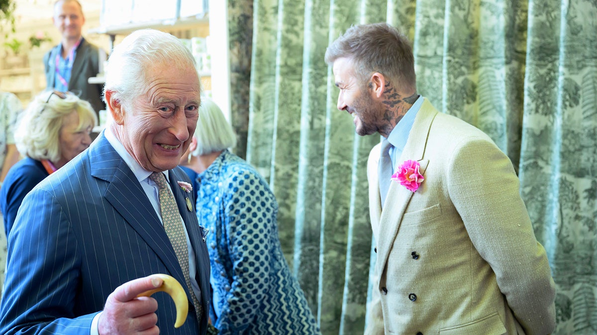 King Charles laughing as David Beckham speaks to someone in a suit with a floral pin.