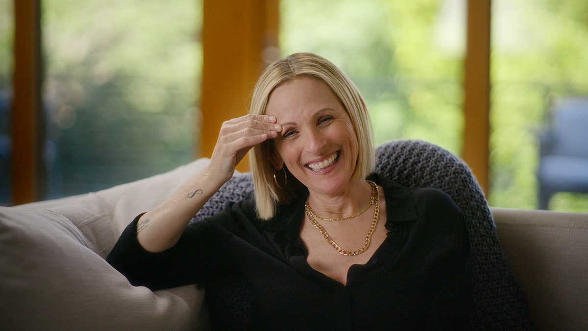 Marlee Matlin smiling wearing a black sweater sitting on a couch.
