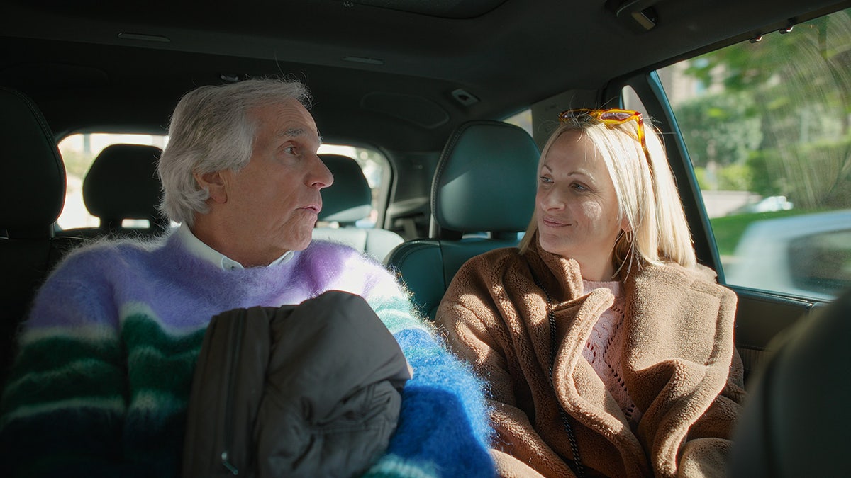 Henry Winkler and Marlee Matlin sitting inside a car and looking at each other.