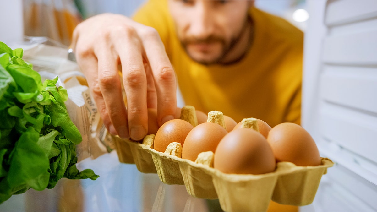 A man grabs an egg as seen from inside the refrigerator.