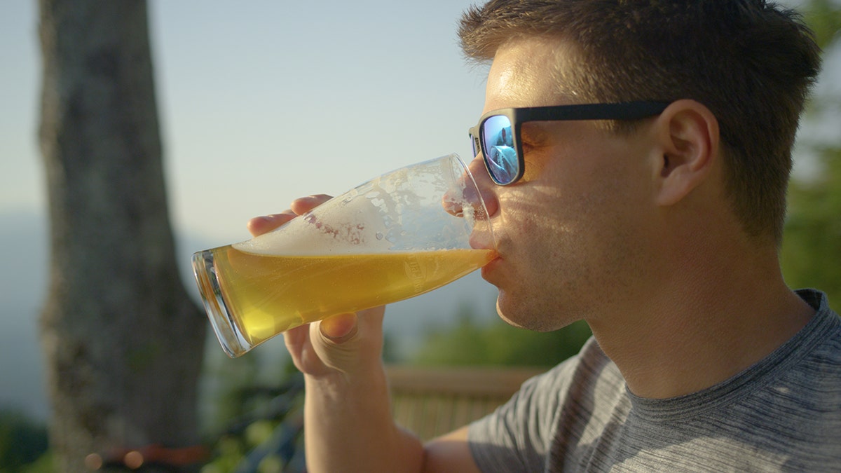 A man wearing sunglasses drinks beer from a glass while outside.
